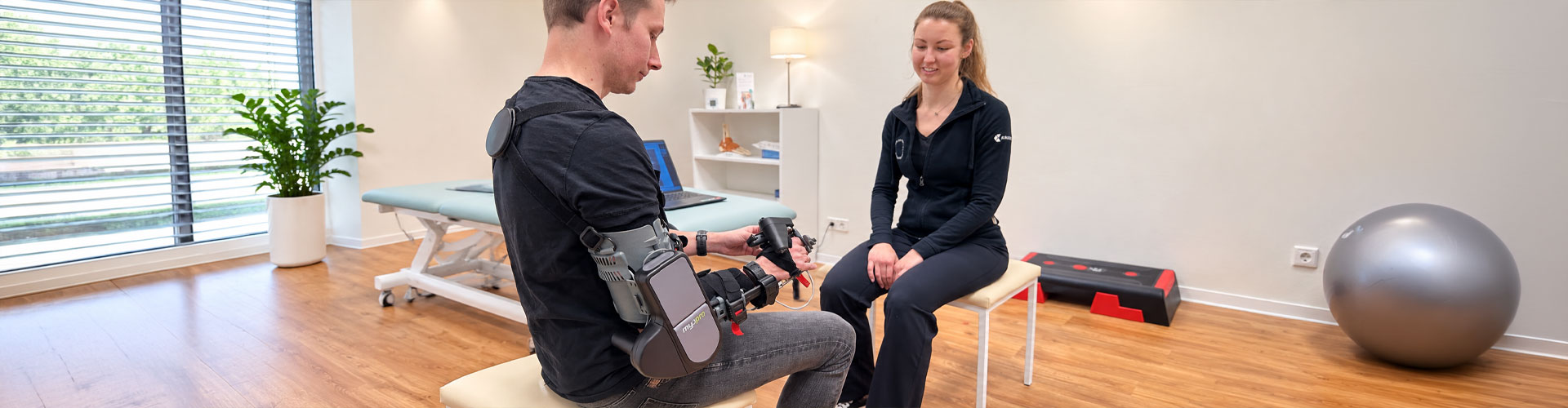 Ein Mann mit MyoPro-Armorthese im Gespräch mit einer Fachkraft in einem Therapieraum.